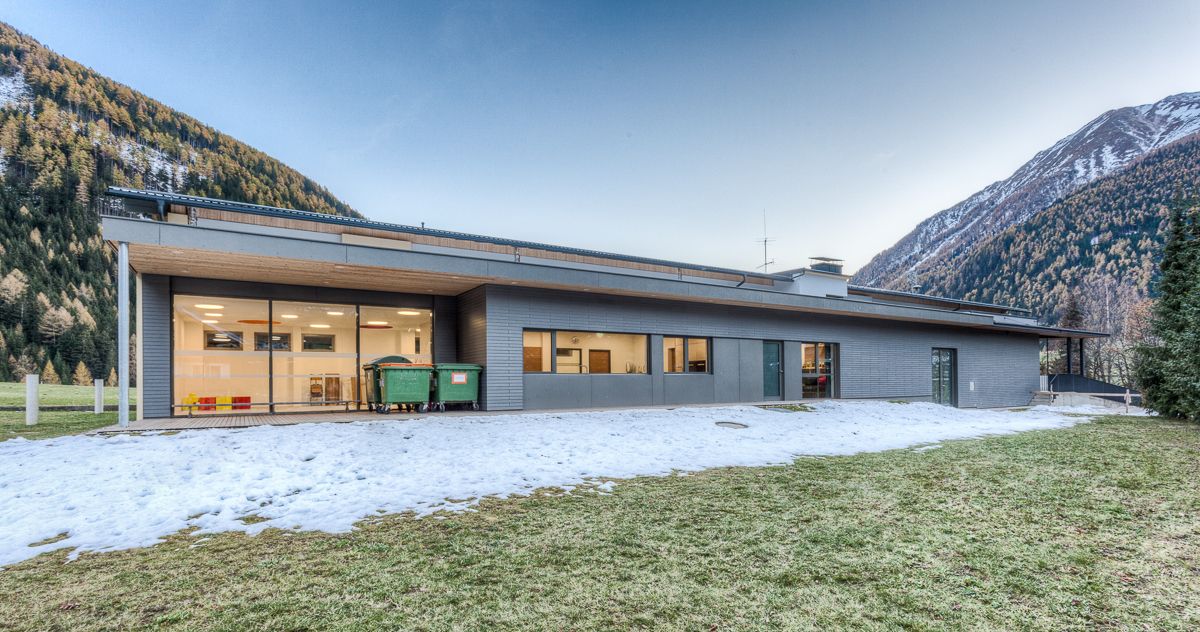 Modernes Gebäude mit großen Fenstern, Recycling-Tonnen im Freien, umgeben von Schnee und Bergen im Hintergrund. Licht am frühen Abend.