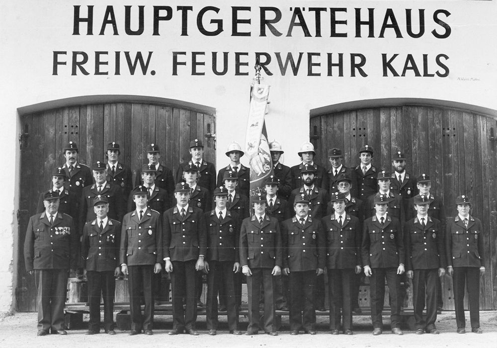 Eine Gruppe von Feuerwehrleuten in Uniform posiert vor dem Hauptgerätehaus in Kals, mit einer Fahne und Holztüren im Rücken.