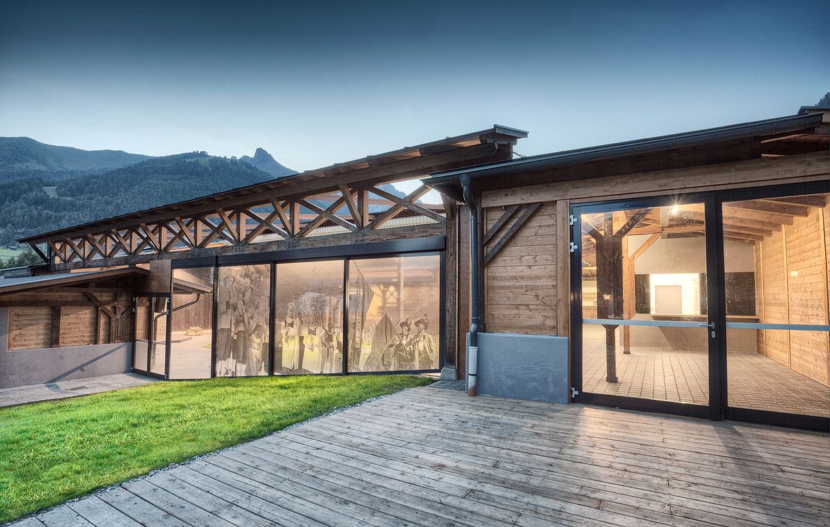 Rustikales Holzgebäude mit großen Glasfenstern, die den Blick auf die malerischen Berge und eine üppige Rasenfläche freigeben, die in der Abenddämmerung beleuchtet wird.