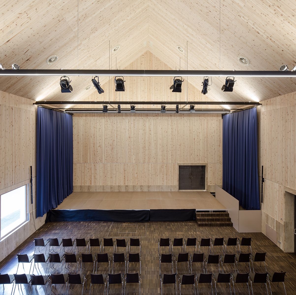 Ein geräumiger hölzerner Hörsaal mit einer Bühne, blauen Vorhängen und Reihen leerer schwarzer Stühle, die von Oberlichtern beleuchtet werden.