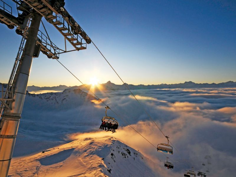 Ein Skilift fährt schneebedeckte Berge hinauf, während die Sonne aufgeht, die Wolken darunter beleuchtet und die Landschaft in ein goldenes Licht taucht.