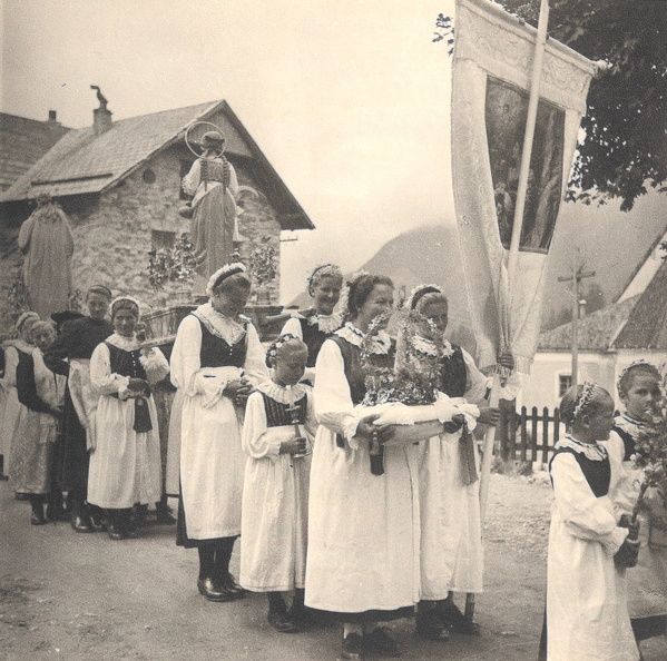 Eine Prozession von Menschen in traditionellen weißen Kleidern trägt Blumen und eine Fahne, im Hintergrund ist eine Statue zu sehen.