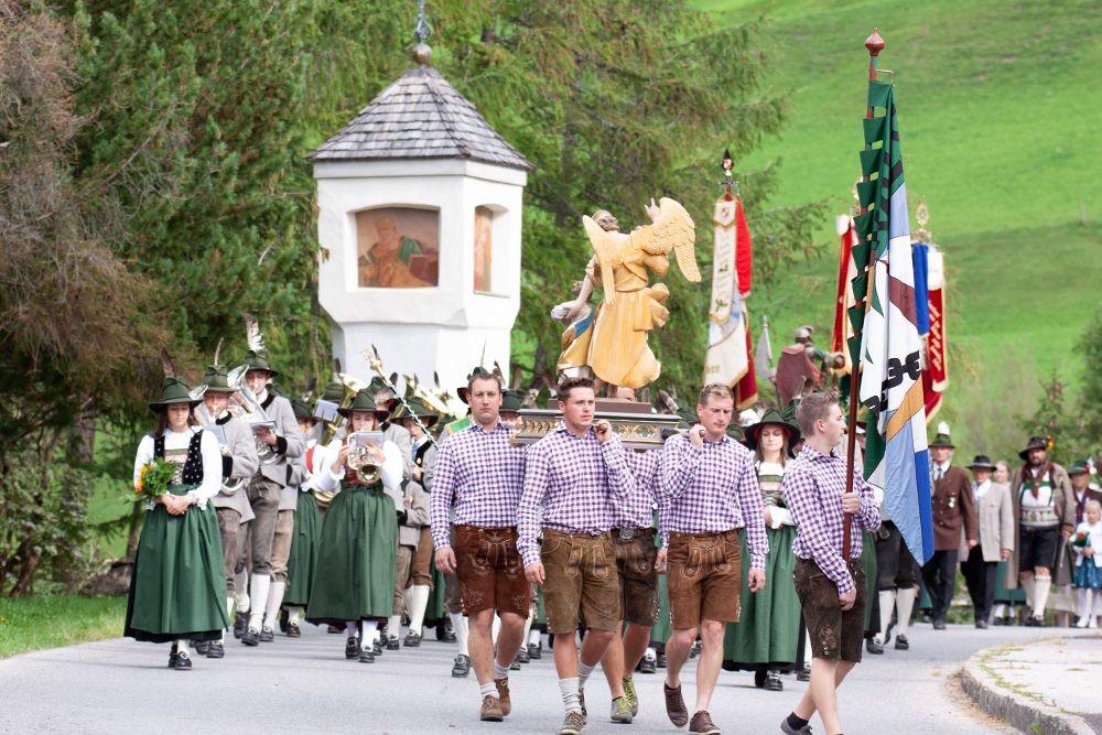Eine festliche Parade mit Menschen in traditioneller Kleidung, die eine Statue und Fahnen tragen, mit einem grünen Hügel im Hintergrund.