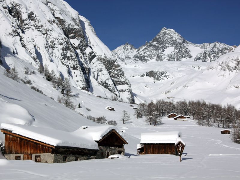Schneebedeckte Almhütten in einem ruhigen Tal, umgeben von majestätischen Bergen unter einem klaren blauen Himmel.