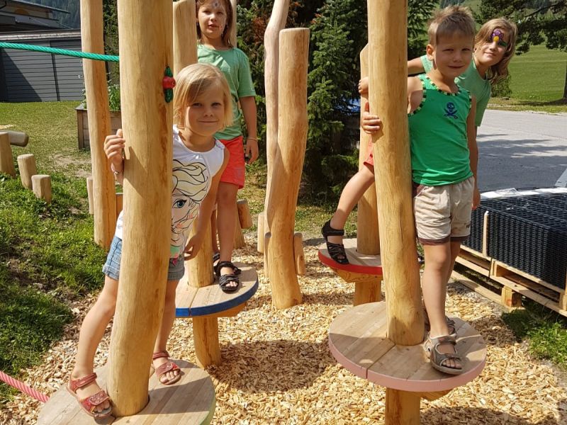 Kinder spielen auf bunten Holzplattformen, die von Baumstämmen gestützt werden, umgeben von Grün und Bergen im Hintergrund.