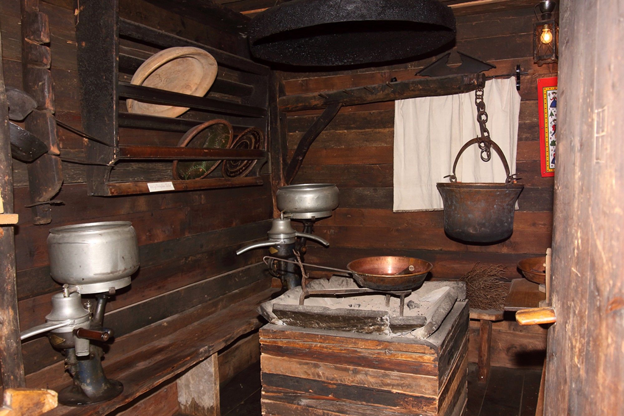 Eine rustikale Holzküche mit alten Kochgeräten, darunter ein Metallherd, ein hängender Kessel und Regale mit Geschirr.