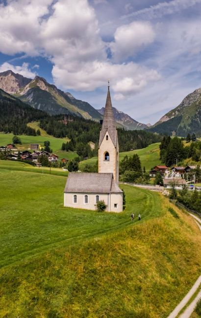 Eine malerische Kirche steht inmitten grüner Felder, umgeben von Bergen und einem Dorf, mit einem gewundenen Weg