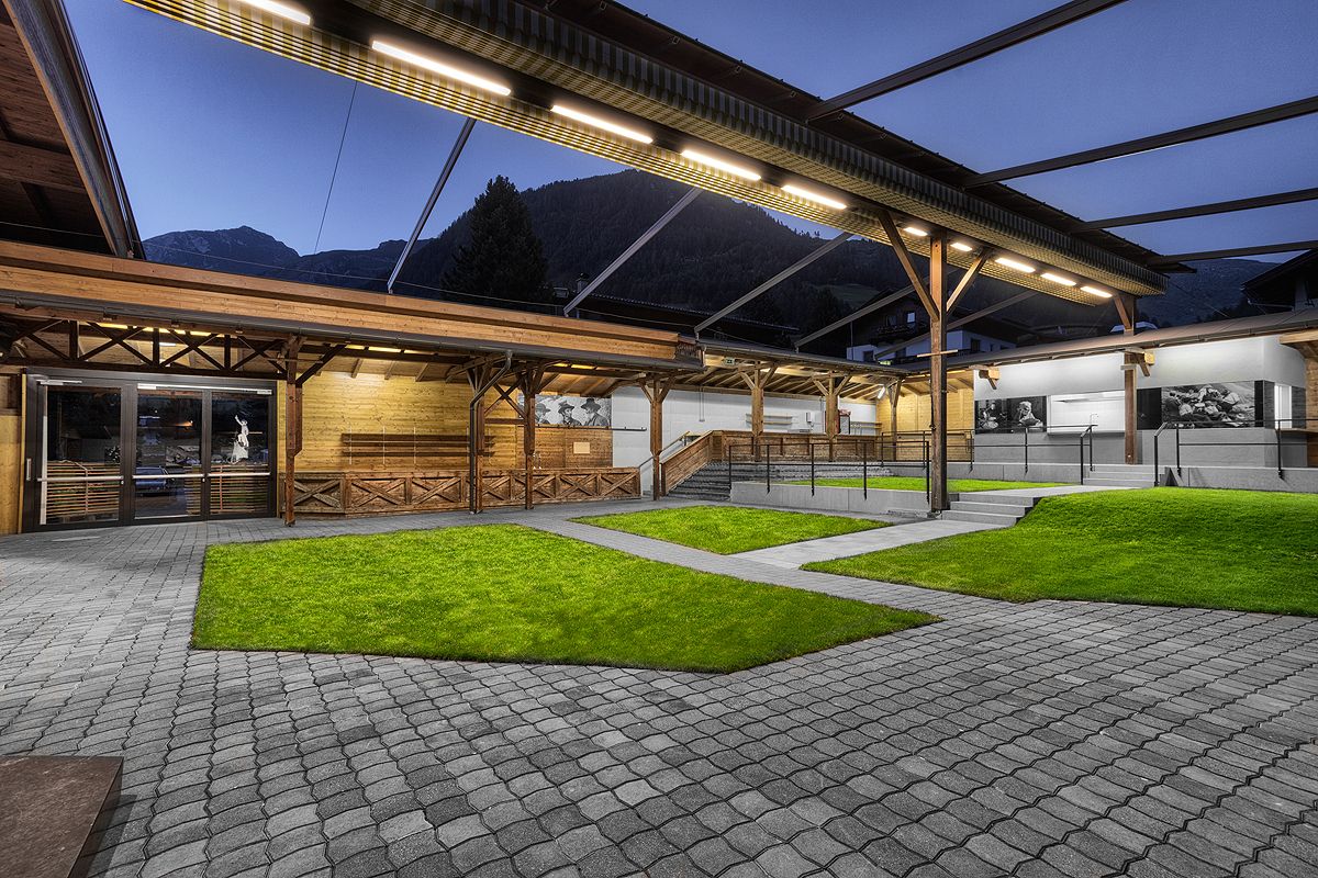 Ein moderner Außenbereich mit hölzernen Strukturen, grünem Gras und Steinwegen, beleuchtet in der Dämmerung vor einer Bergkulisse.