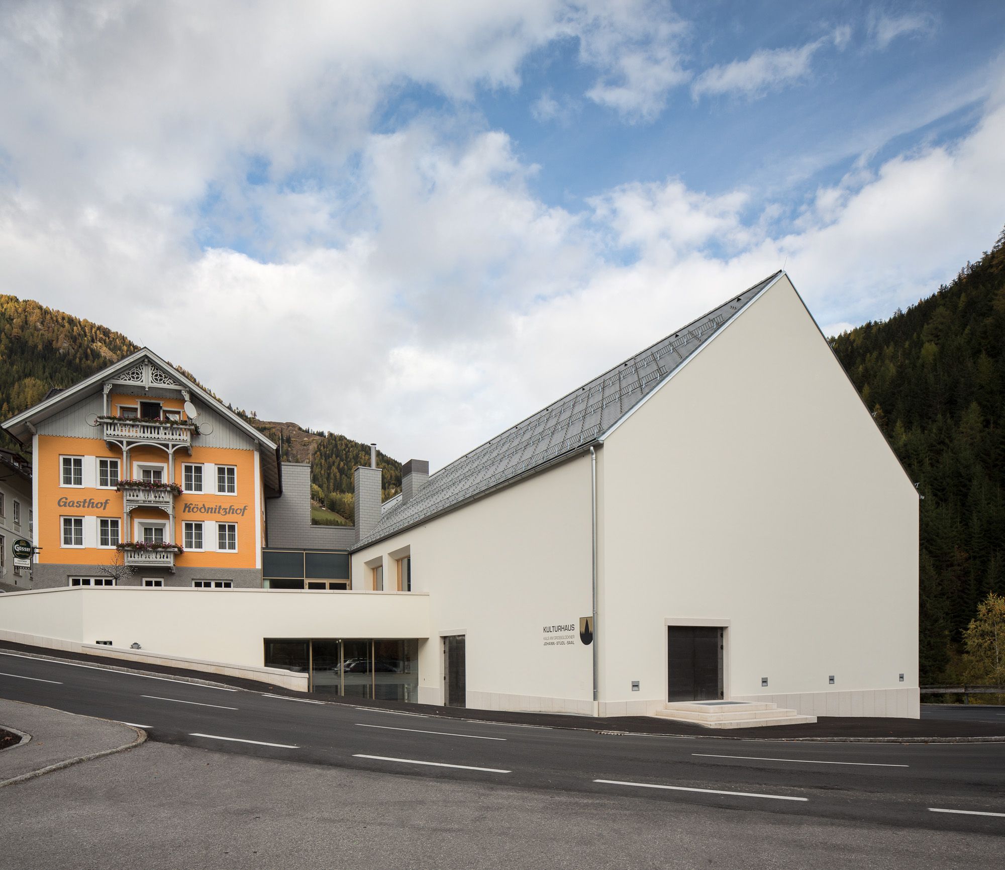 Ein traditioneller orangefarbener Gasthof kontrastiert mit einem modernen weißen Kulturzentrum vor der Kulisse der Berge und des bewölkten Himmels.