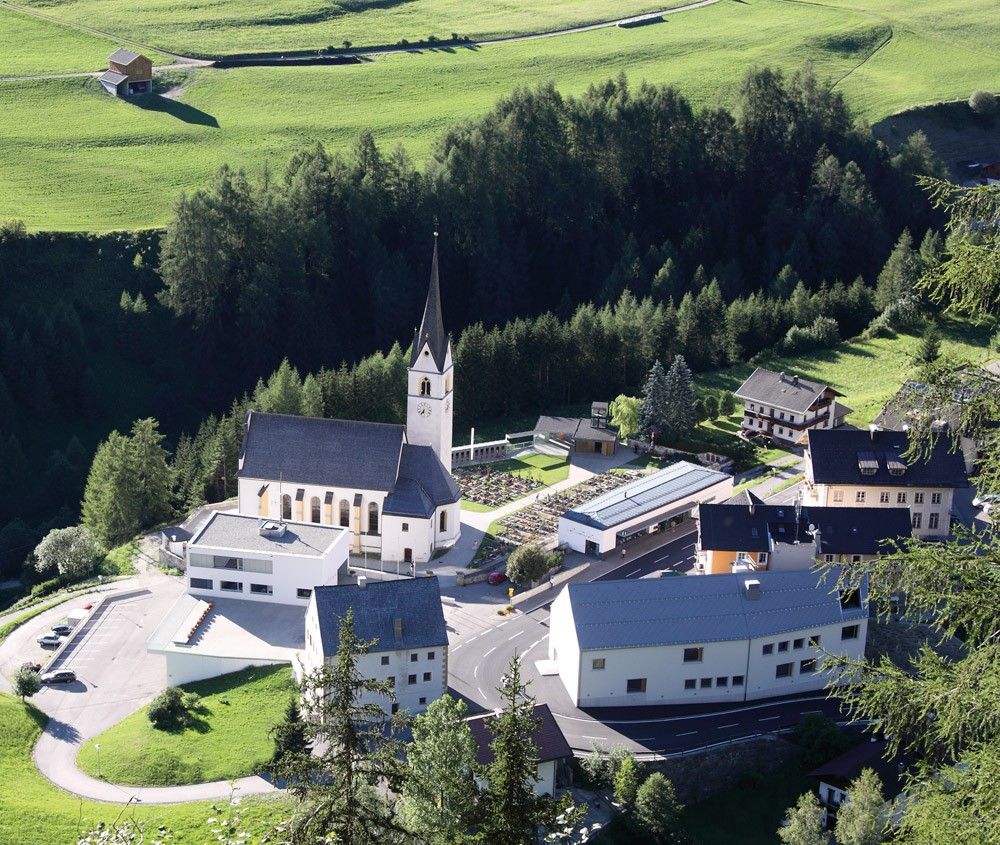 Luftaufnahme eines malerischen Dorfes mit einer Kirchturmuhr, umliegenden Grünflächen und nahe gelegenen Bauernhöfen inmitten der Berge.