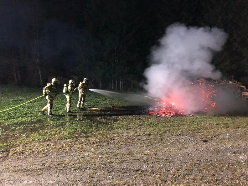 Feuerwehrleute löschen nachts ein glühendes Feuer, wobei Rauch aufsteigt und Schutzkleidung vor einem dunklen Waldhintergrund zu sehen ist.