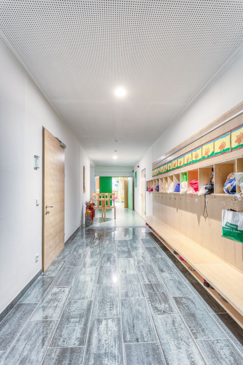 Ein heller, moderner Flur mit gefliestem Boden, einer Holztür, Ablagefächern für Taschen und einem grünen Klassenzimmereingang am Ende.