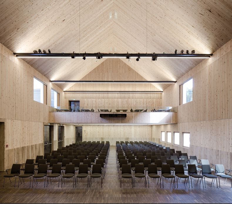 Das Innere eines geräumigen Holzsaals mit Stuhlreihen, großen Fenstern und einem Balkon ist ideal für Veranstaltungen und Tagungen.