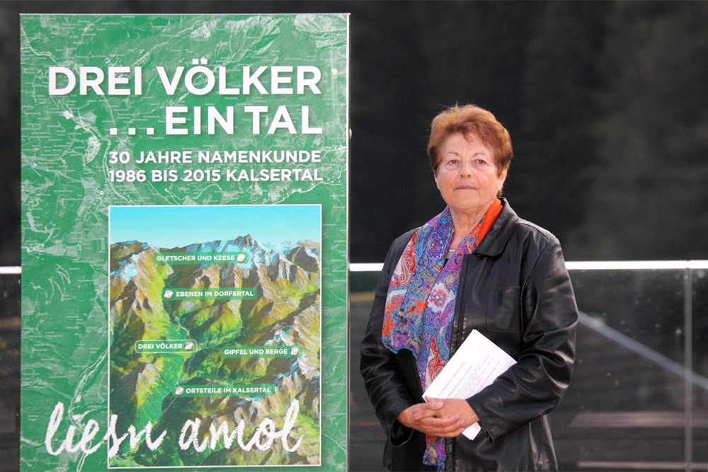 Eine Frau mit einem bunten Schal steht neben einem grünen Plakat mit dem Titel „Drei Völker ... ein Tal“, das 30 Jahre Kalsertal feiert.