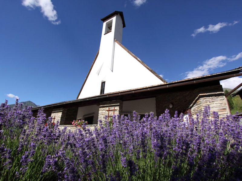 Eine bezaubernde weiße Kirche mit einem hohen Glockenturm, umgeben von leuchtenden lavendelfarbenen Blumen vor einem strahlend blauen Himmel.