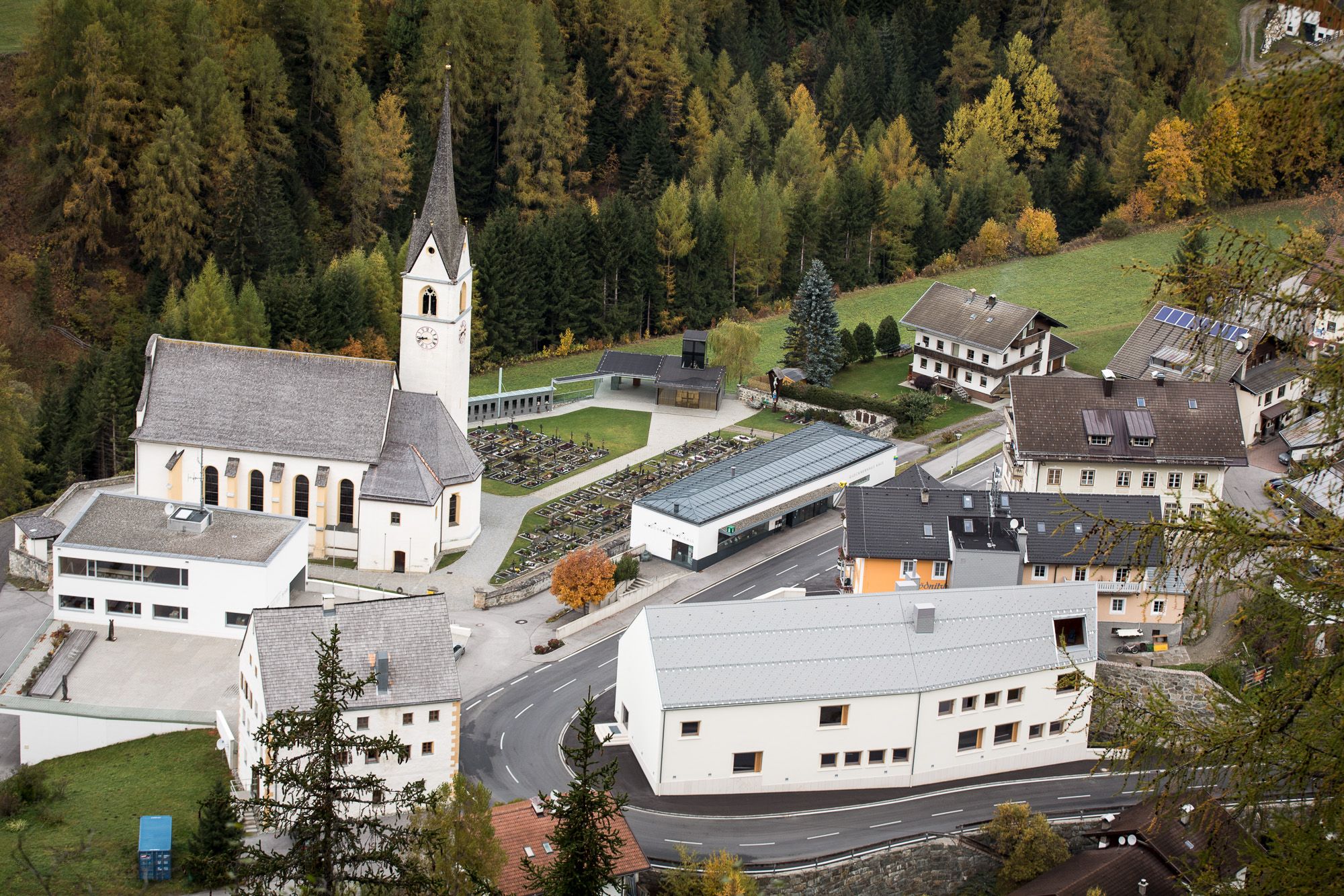Luftaufnahme eines malerischen Dorfes mit einer Kirche, einem Friedhof und umliegenden Häusern inmitten herbstlich gefärbter Bäume.