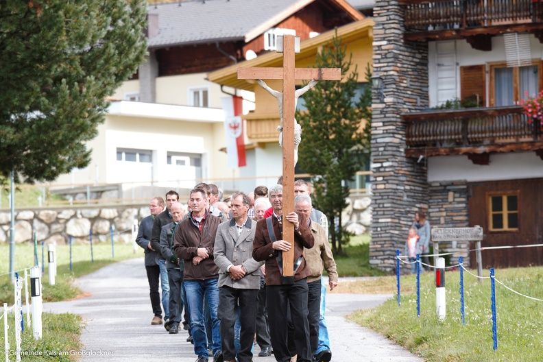 Eine Gruppe von Menschen geht einen Weg entlang, wobei eine Person ein großes Holzkreuz vor einer Kulisse von Gebäuden und Bäumen trägt.