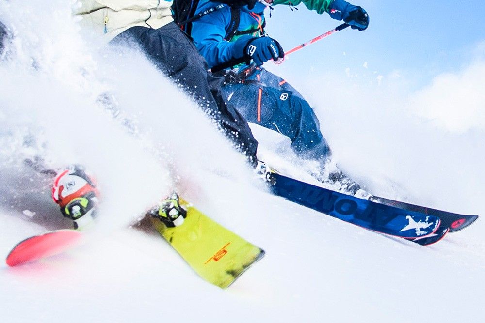 Zwei Skifahrer fahren durch den frischen Pulverschnee und präsentieren ihre Skikleidung und ihre Action vor einem strahlend blauen Himmel.
