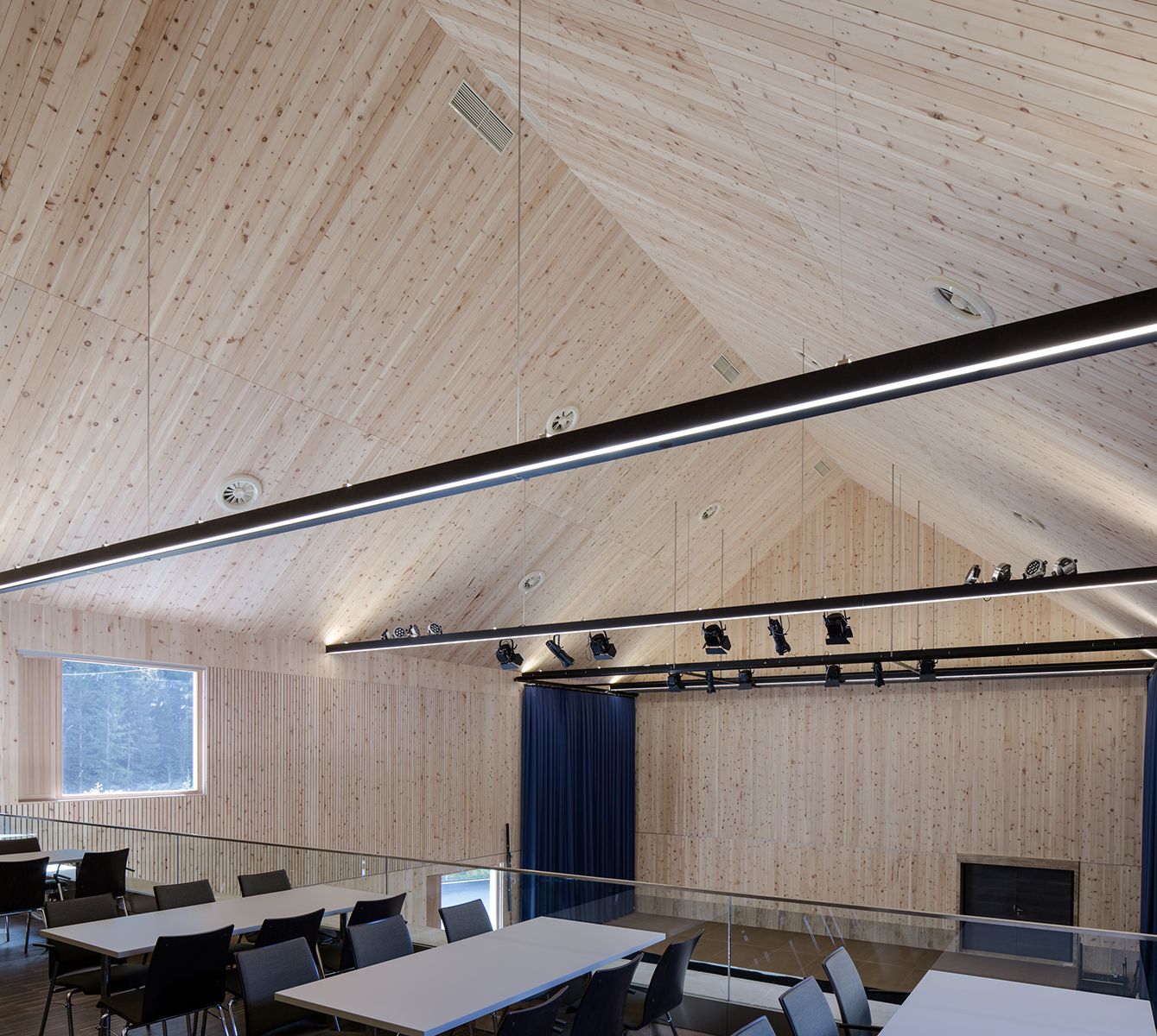 Modernes Holzinterieur in einem geräumigen Mehrzweckraum mit hohen Decken, Schienenbeleuchtung und Tischen für Zusammenkünfte.