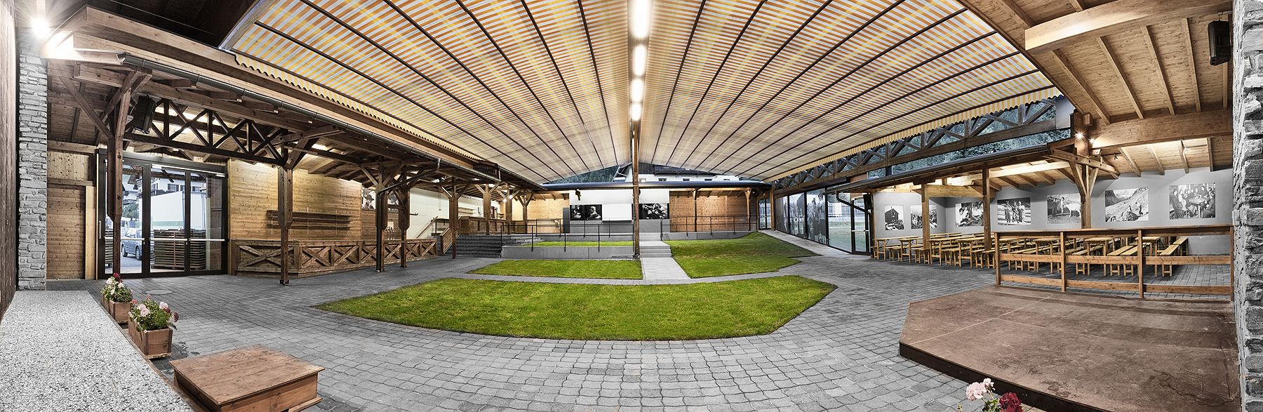 Ein geräumiger, moderner Veranstaltungsort unter freiem Himmel mit Holzstrukturen, grünem Gras und Essbereichen mit großen Fenstern und Fotos an den Wänden.