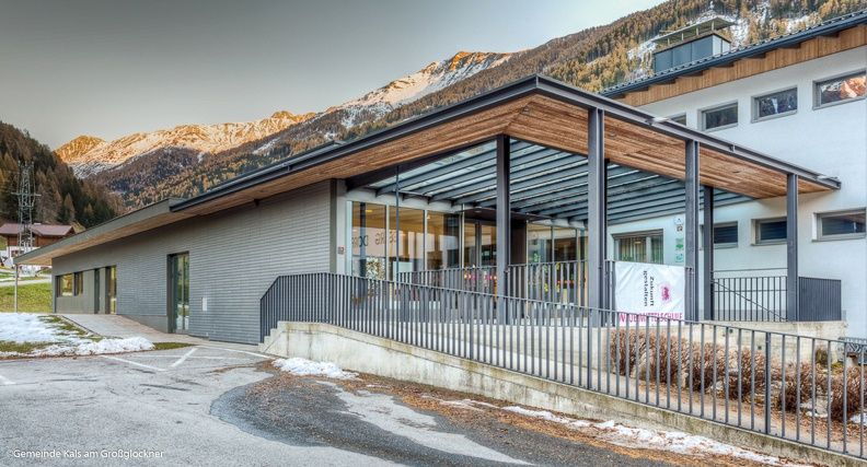 Modernes Gebäude mit Holzüberdachung und großem Glaseingang vor der Kulisse schneebedeckter Berge.
