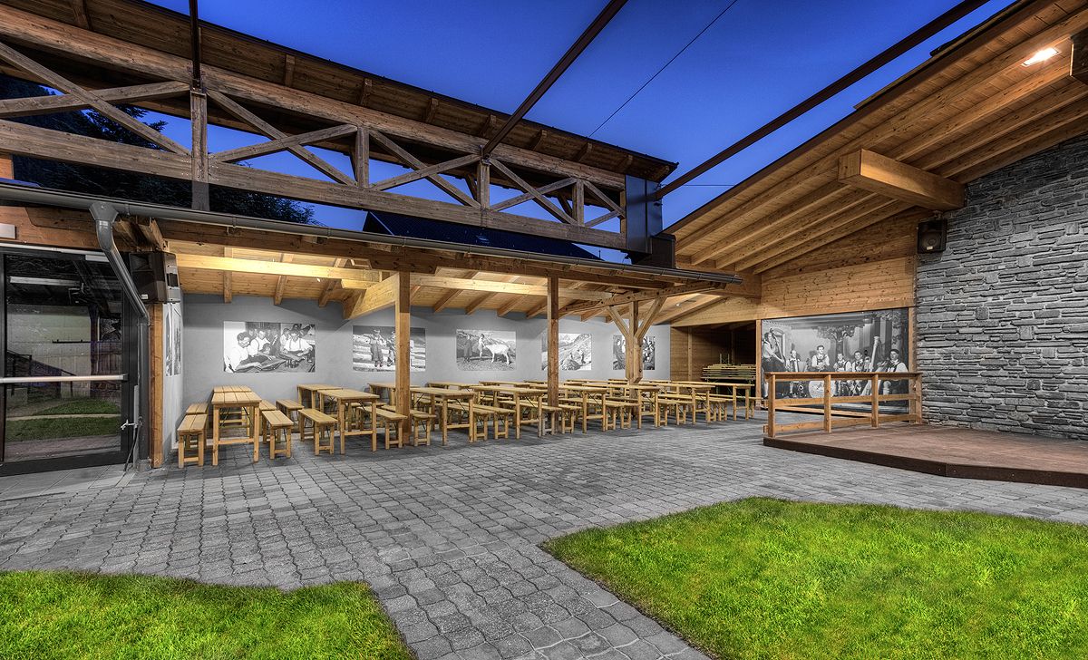 Essbereich im Freien mit Holztischen, einem hohen Dach und historischen Fotografien an der Wand, die in der Dämmerung beleuchtet werden.