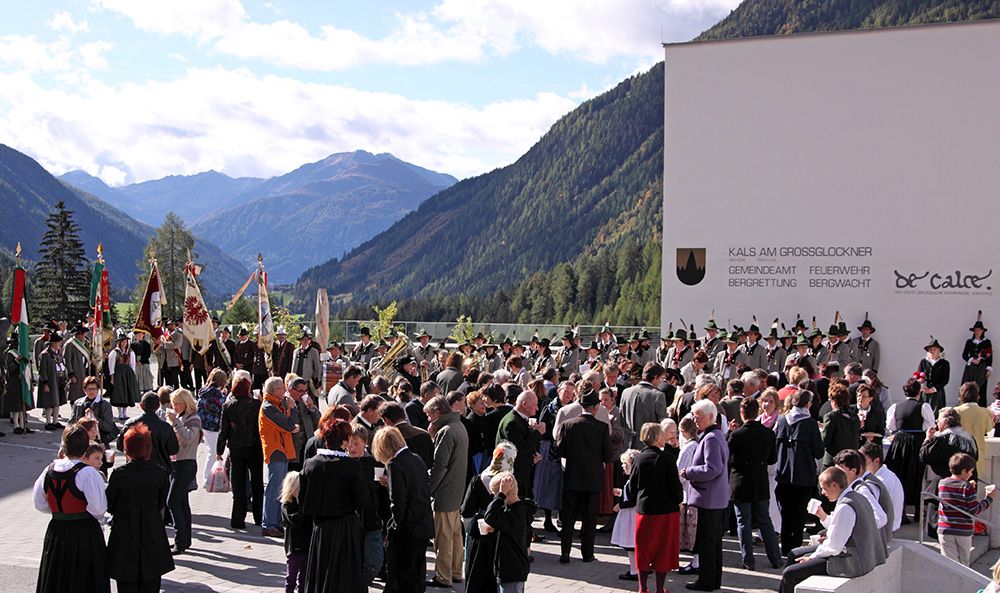 Eine lebhafte Versammlung in traditioneller Kleidung findet unter freiem Himmel statt, mit den Bergen im Hintergrund und einem Gebäude, das lokale Namen und Symbole trägt.