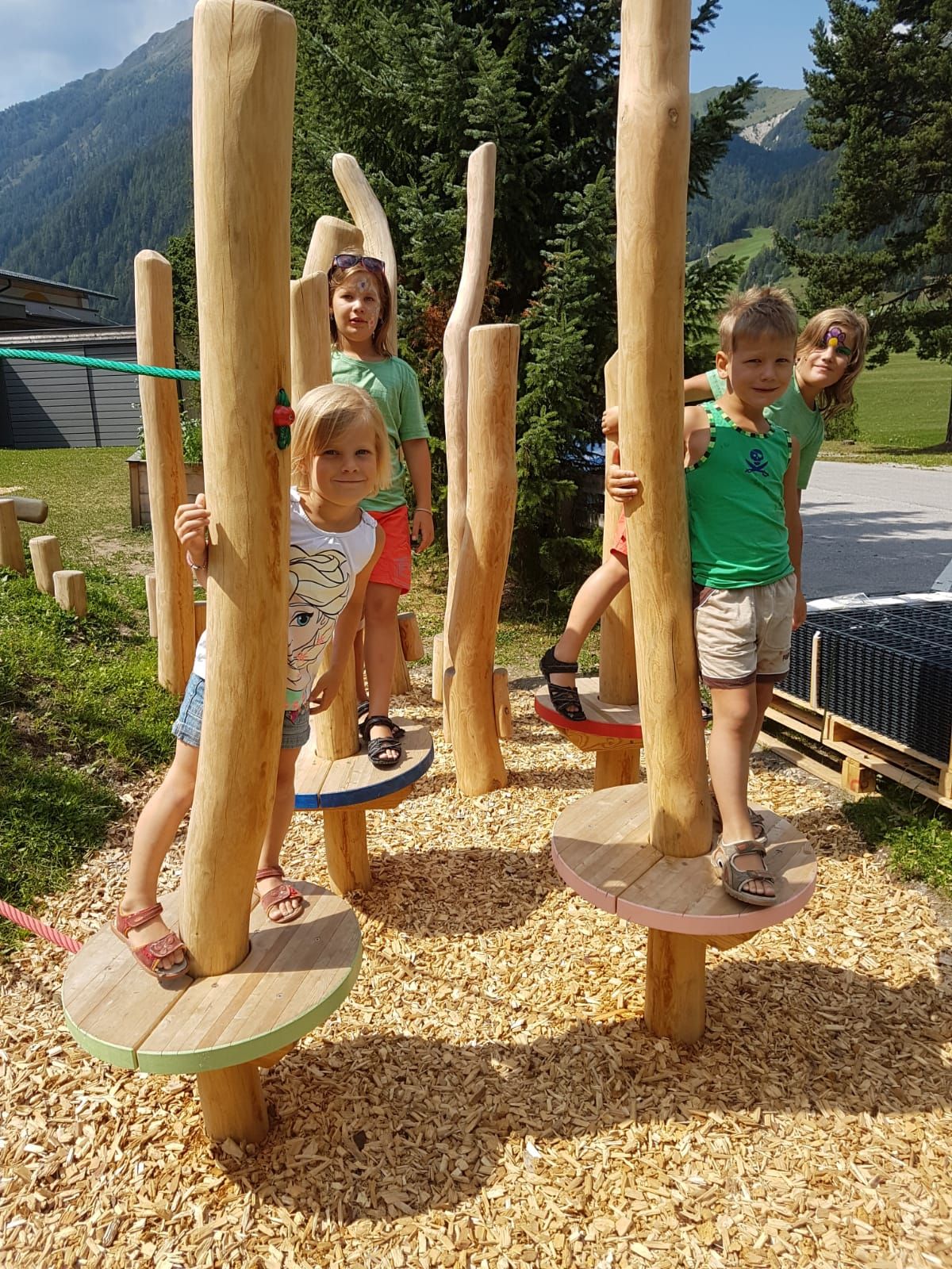 Kinder spielen auf bunten Holzplattformen, die von Baumstämmen gestützt werden, umgeben von Grün und Bergen im Hintergrund.