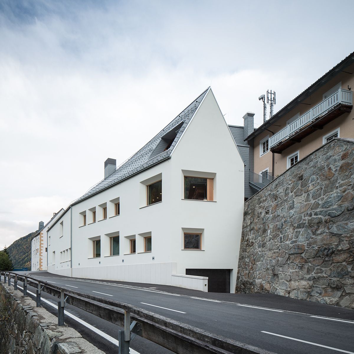 Modernes weißes Haus mit einem einzigartigen Schrägdach, angrenzend an eine Steinmauer und ältere Gebäude, vor einem bewölkten Himmel.