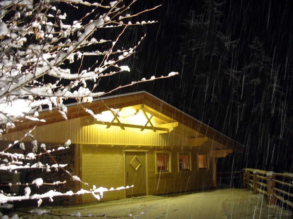 Eine gemütliche Holzhütte, die in einer verschneiten Nacht beleuchtet wird, umgeben von Bäumen, während die Schneeflocken sanft in die Luft fallen.