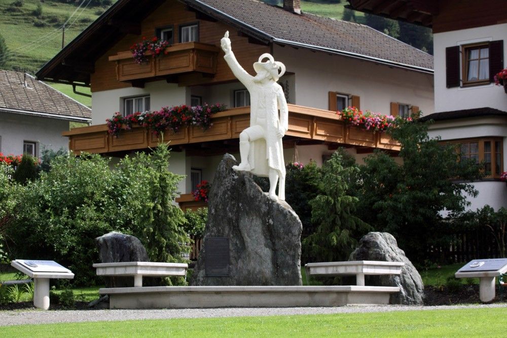 Eine weiße Statue eines nach oben zeigenden Mannes steht auf einem Felsen, umgeben von Grün und traditionellen Holzhäusern mit Blumenkästen.