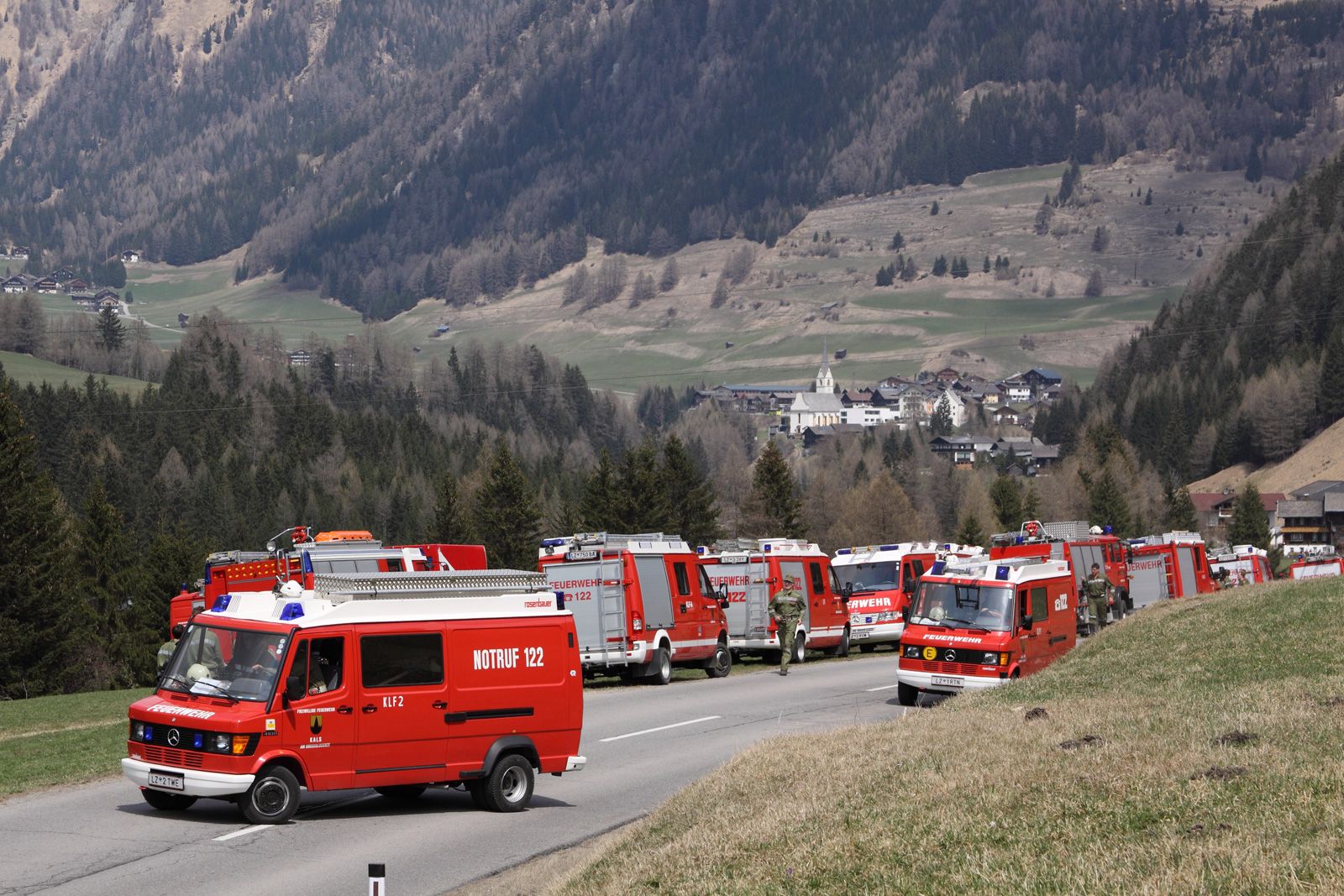 Ein Konvoi roter Einsatzfahrzeuge, darunter Feuerwehrfahrzeuge und Lieferwagen, reiht sich auf einer Landstraße vor einer Bergkulisse auf.