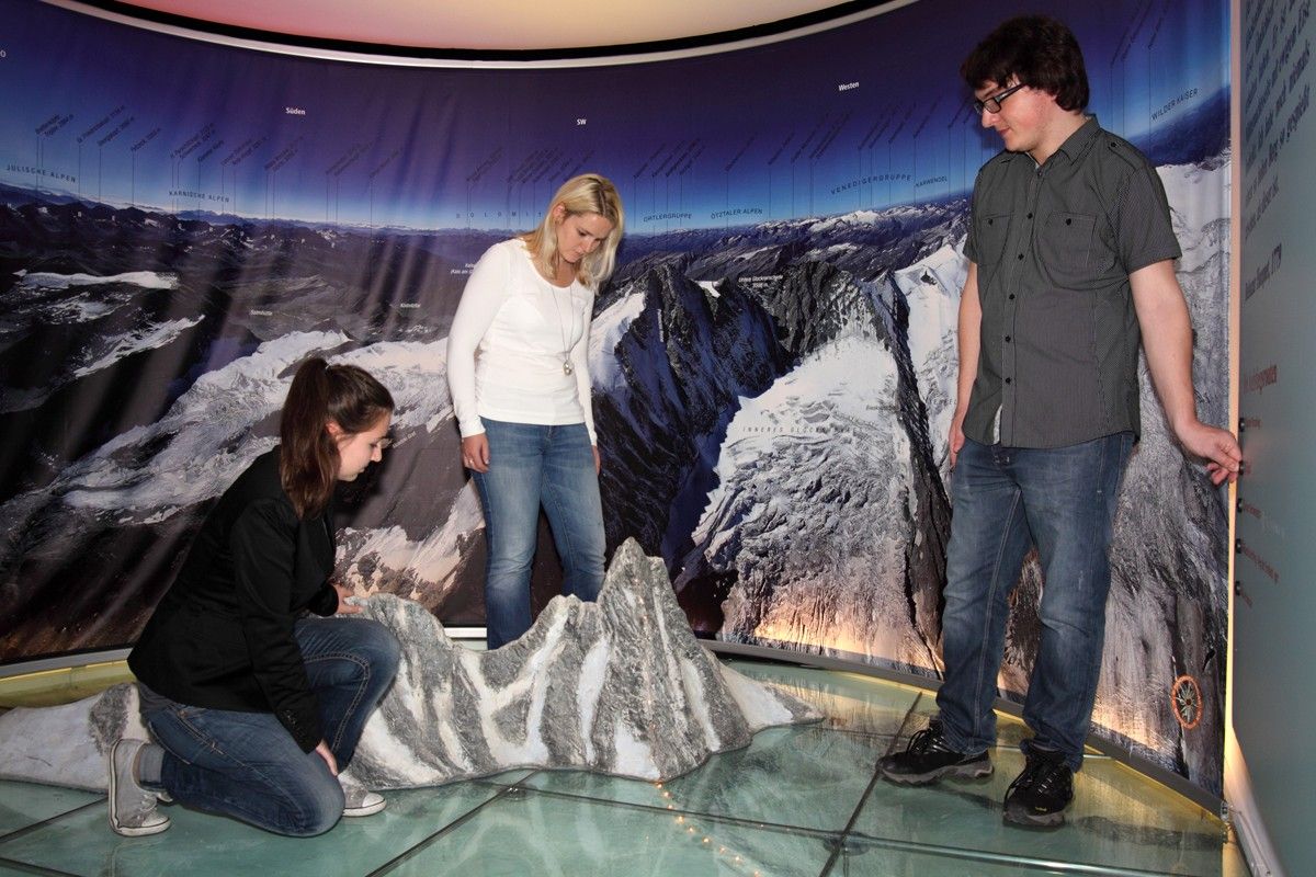 Zwei Bergsteiger bewegen sich auf einem schneebedeckten Grat am Kleinglockner, umgeben von riesigen Bergketten und einem Wolkenmeer unter ihnen. Schwarz-Weiß-Foto.