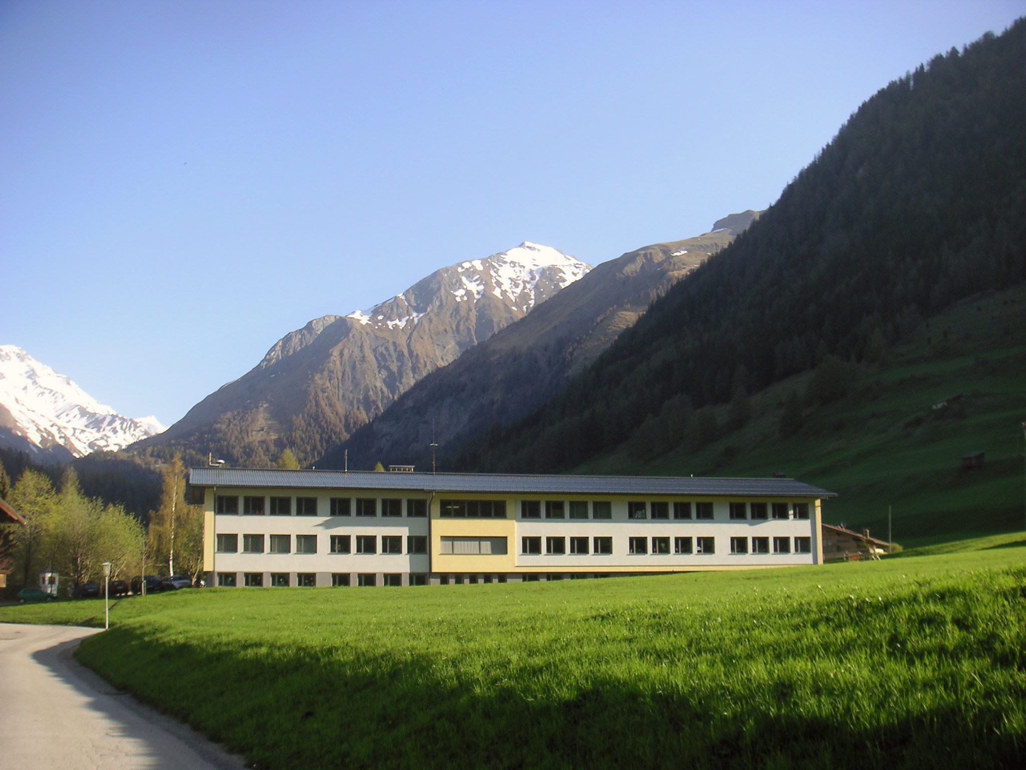 Ein modernes Gebäude mit großen Fenstern steht auf einer grünen Wiese, umgeben von majestätischen schneebedeckten Bergen unter einem klaren blauen Himmel.