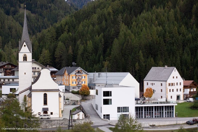 Eine malerische Dorfansicht mit einer Kirche mit hohem Kirchturm, traditionellen und modernen Gebäuden vor der Kulisse üppiger Berge.