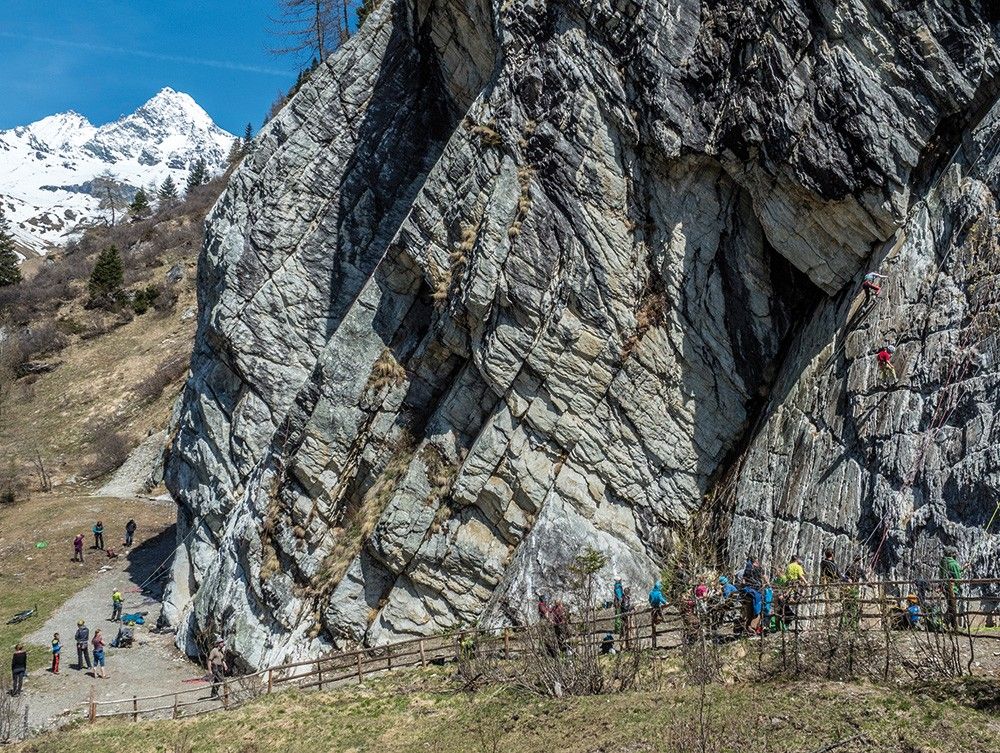 Kletterer bezwingen eine steile Felswand in einer landschaftlich reizvollen Berggegend, während die Zuschauer an einem sonnigen Tag in der Nähe zuschauen.