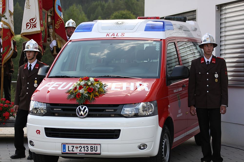Ein mit Blumen geschmückter rot-weißer Feuerwehrauto, flankiert von zwei Uniformierten, bei einer Zeremonie im Freien.
