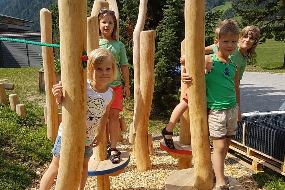 Kinder spielen auf einem hölzernen Klettergerüst in einem sonnigen Park, umgeben von Bäumen und Bergen, und genießen ein Abenteuer im Freien.