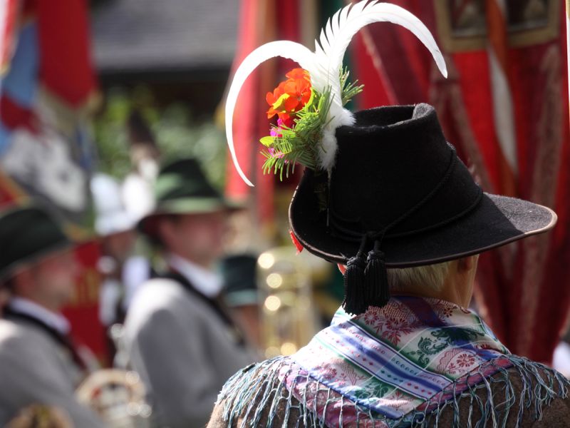 Eine Person trägt bei einer festlichen Veranstaltung einen traditionellen schwarzen Hut, der mit einer Feder und Blumen geschmückt ist, und einen bunten Schal um die Schultern.