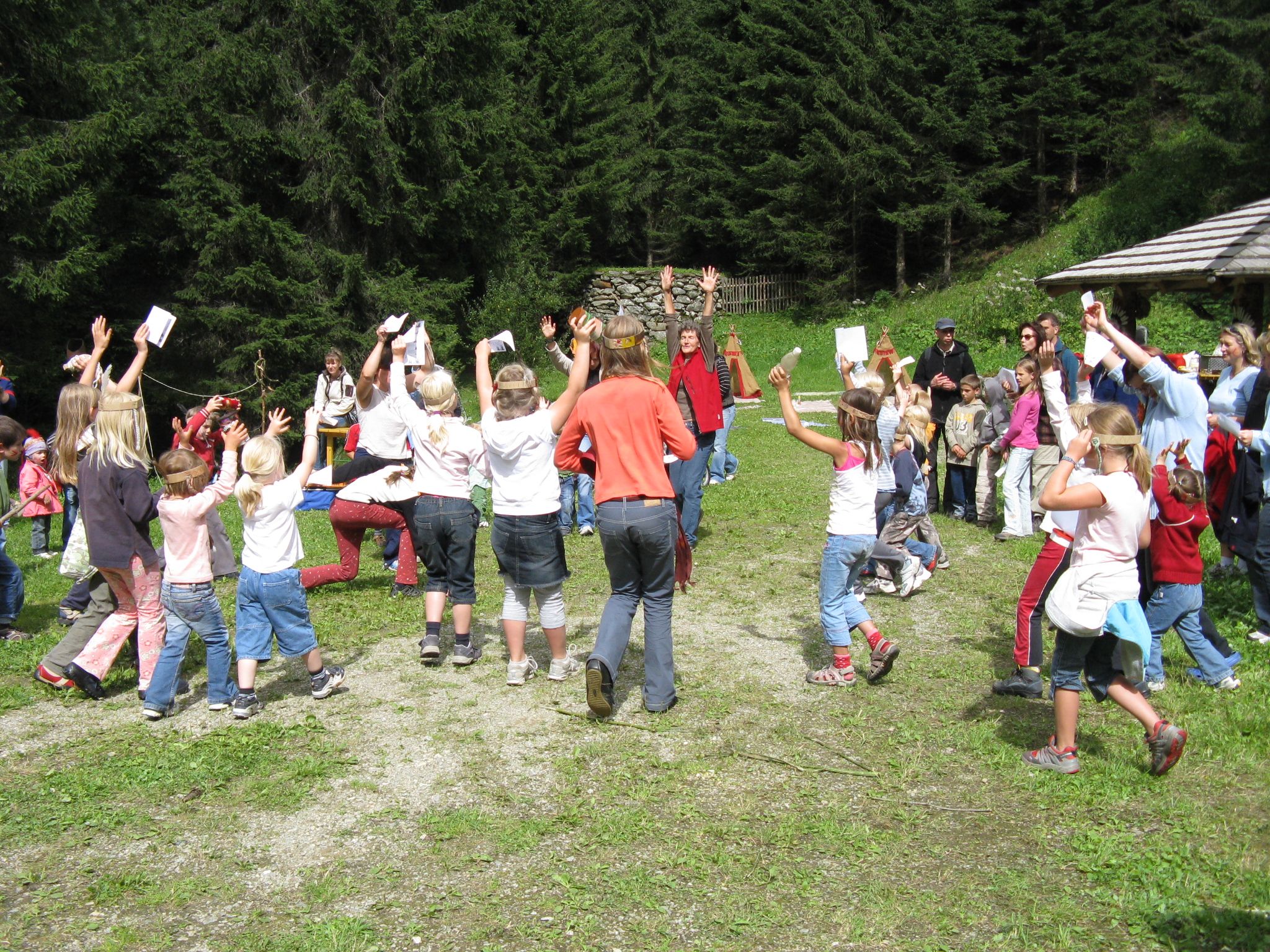 Eine lebhafte Gruppe von Kindern tanzt und feiert mit erhobenen Händen inmitten einer grünen Waldkulisse im Freien.