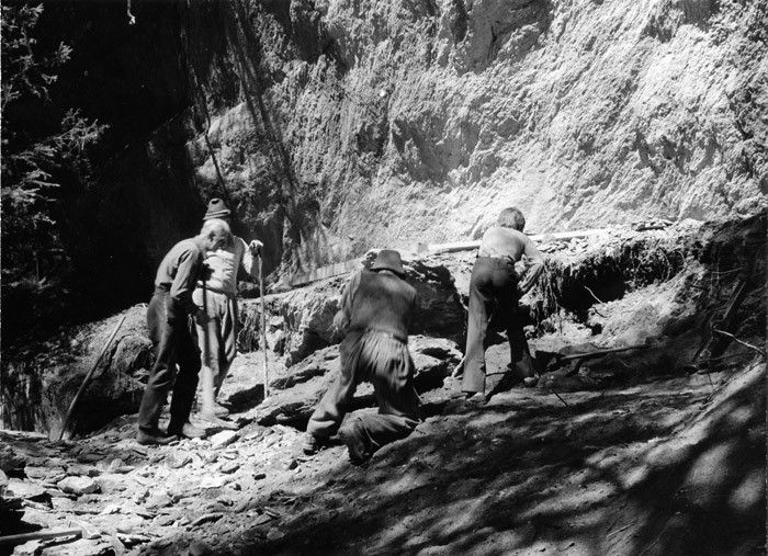 Vier Männer in historischer Kleidung bei der Arbeit mit Werkzeugen in einer rauen, bewaldeten Gegend, die auf frühe Ausgrabungsaktivitäten hindeutet.