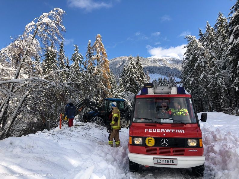 Feuerwehrleute helfen einem schneebedeckten Fahrzeug in einer Winterlandschaft, mit schneebedeckten Bäumen und Bergen im Hintergrund.