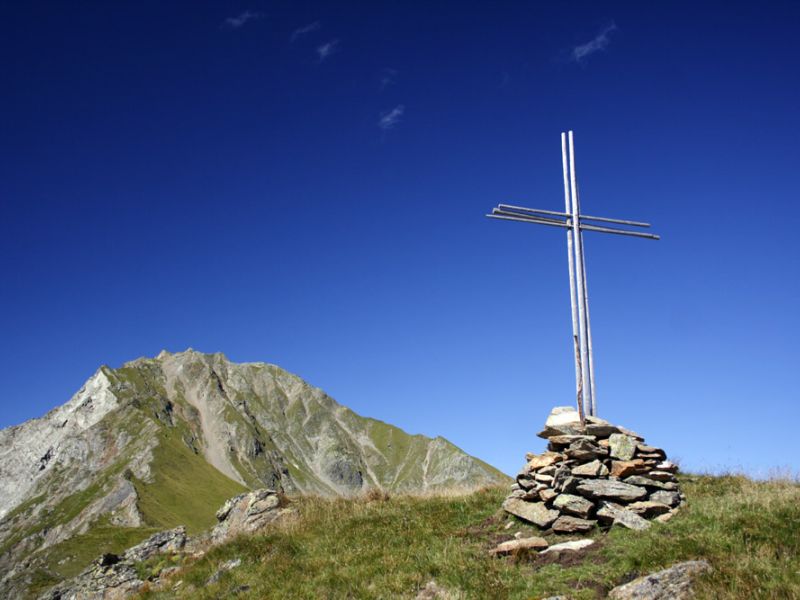 Ein Metallkreuz auf einem Steinhügel, mit einem klaren blauen Himmel und schroffen Gipfeln im Hintergrund.