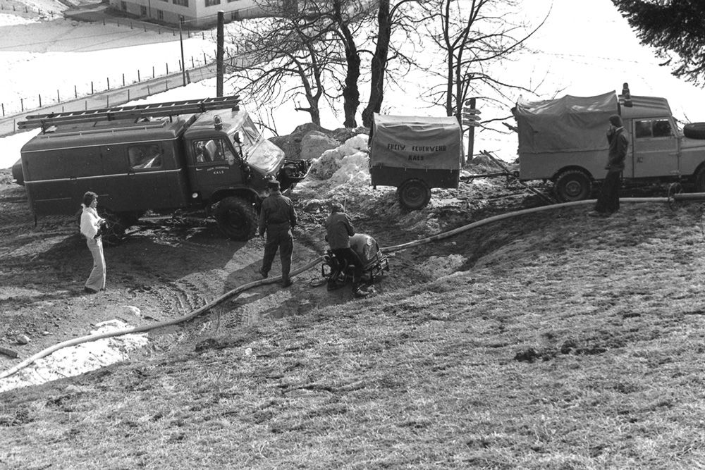 Ein altes Schwarz-Weiß-Foto von Rettungskräften, die mit Fahrzeugen und Schläuchen in einer verschneiten Landschaft arbeiten und bei einem Notfalleinsatz helfen.