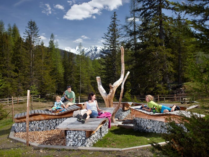 Eine Familie entspannt sich auf natürlichen Holzbänken, umgeben von Bäumen und Bergen unter einem blauen Himmel.