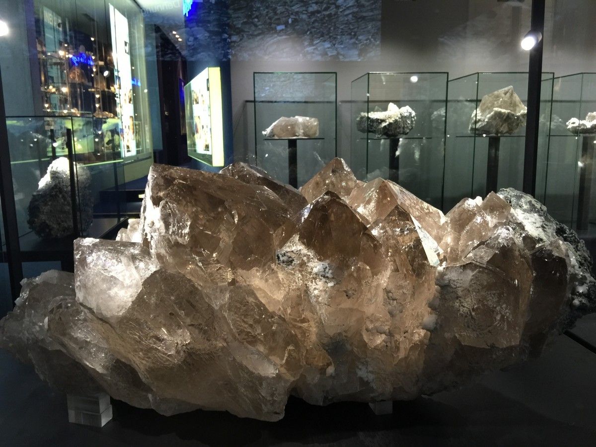 Ein großer Kristallcluster mit funkelnden Facetten, der in einem Museum ausgestellt ist, umgeben von kleineren Mineralien in Vitrinen.