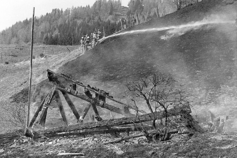 Feuerwehrleute löschen einen schwelenden Hang mit verkohlten Holzresten und verbrannter Erde in einer trostlosen, bewaldeten Landschaft.