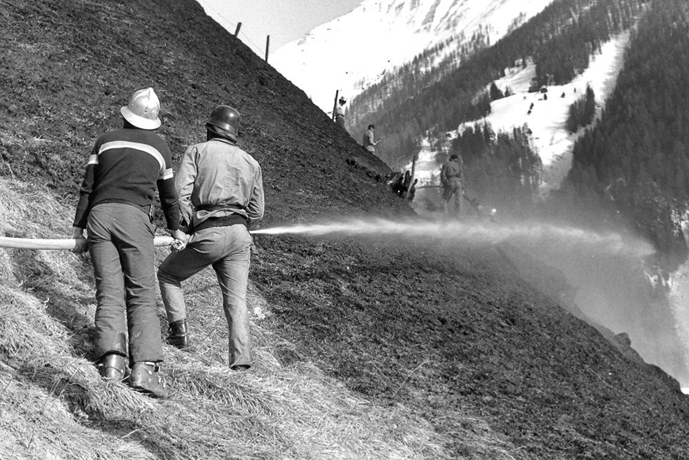 Zwei Feuerwehrleute versprühen Wasser auf einem abschüssigen, trockenen Gelände, um einen Waldbrand zu bekämpfen, mit schneebedeckten Bergen und anderen Feuerwehrleuten im Hintergrund.