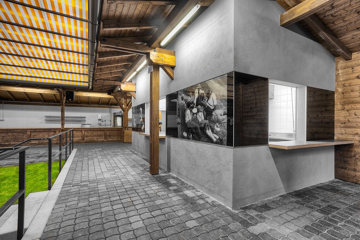 Moderner Außenbereich mit einem Kiosk mit grauen Wänden und historischen Schwarz-Weiß-Fotos, umgeben von Holzstrukturen und Kopfsteinpflasterwegen.