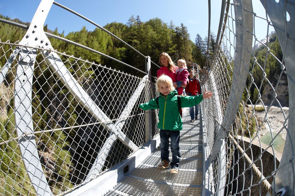 Eine Gruppe von Kindern und Erwachsenen, die auf einer Hängebrücke spazieren gehen, umgeben von üppigen grünen Bäumen und einer felsigen Schlucht unter ihnen.